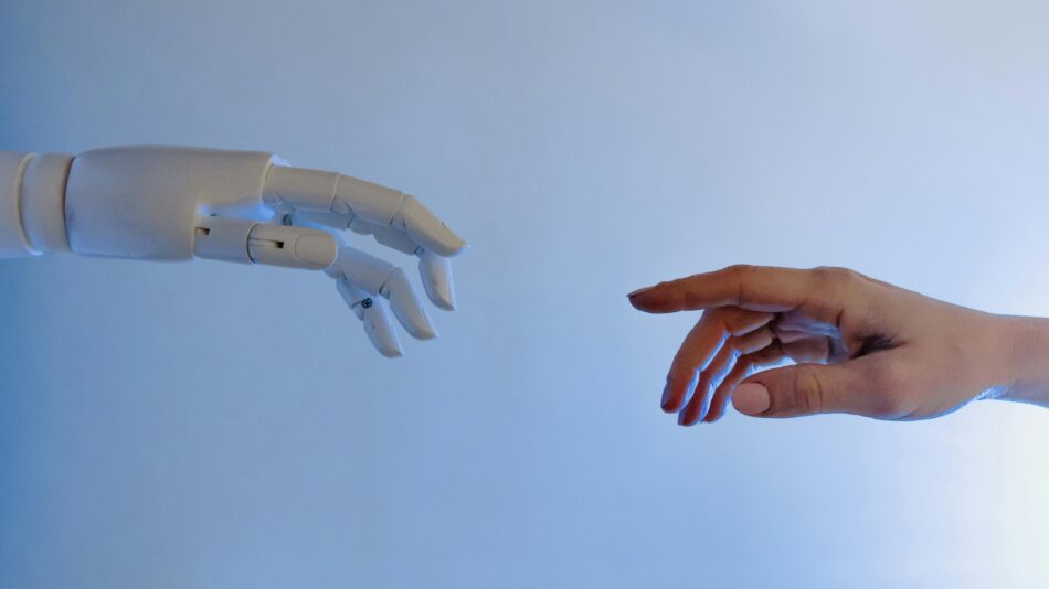 Human hand and robot hand reaching toward each other against a blue sky, symbolising the relationship between people and AI technology