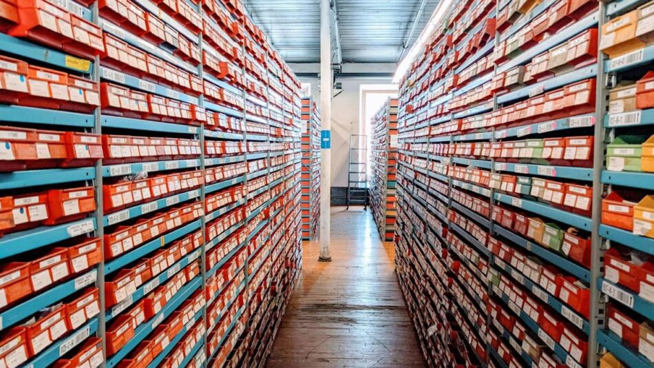 Organised red shoe boxes on metal warehouse shelves, viewed down a narrow aisle