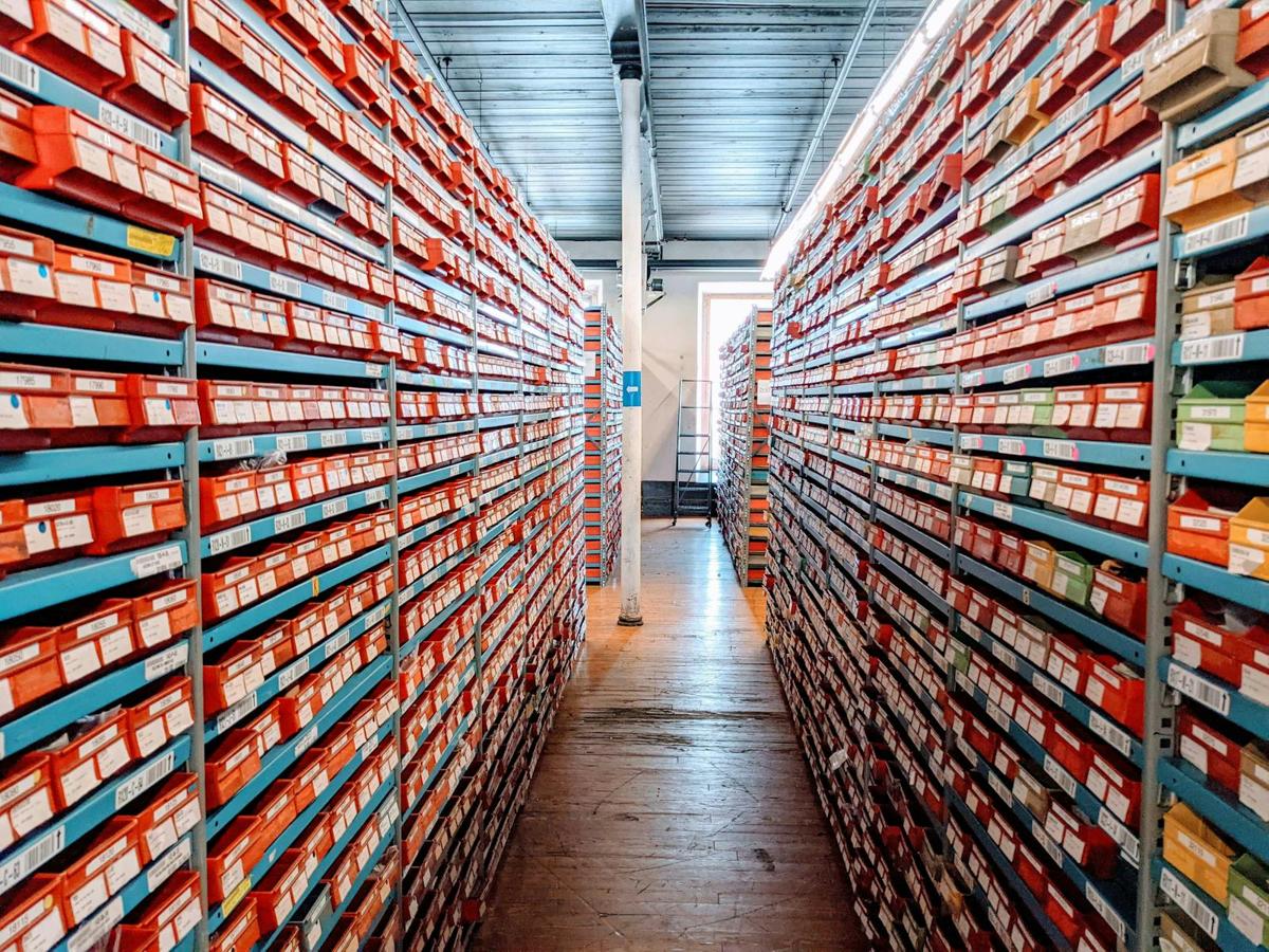 Organised red shoe boxes on metal warehouse shelves, viewed down a narrow aisle